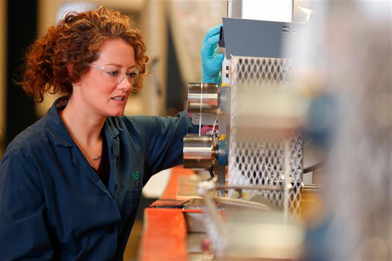 a woman with curly hair, lab safety glasses, aqua gloves, and a blue lab coat stands to the left of a large machine