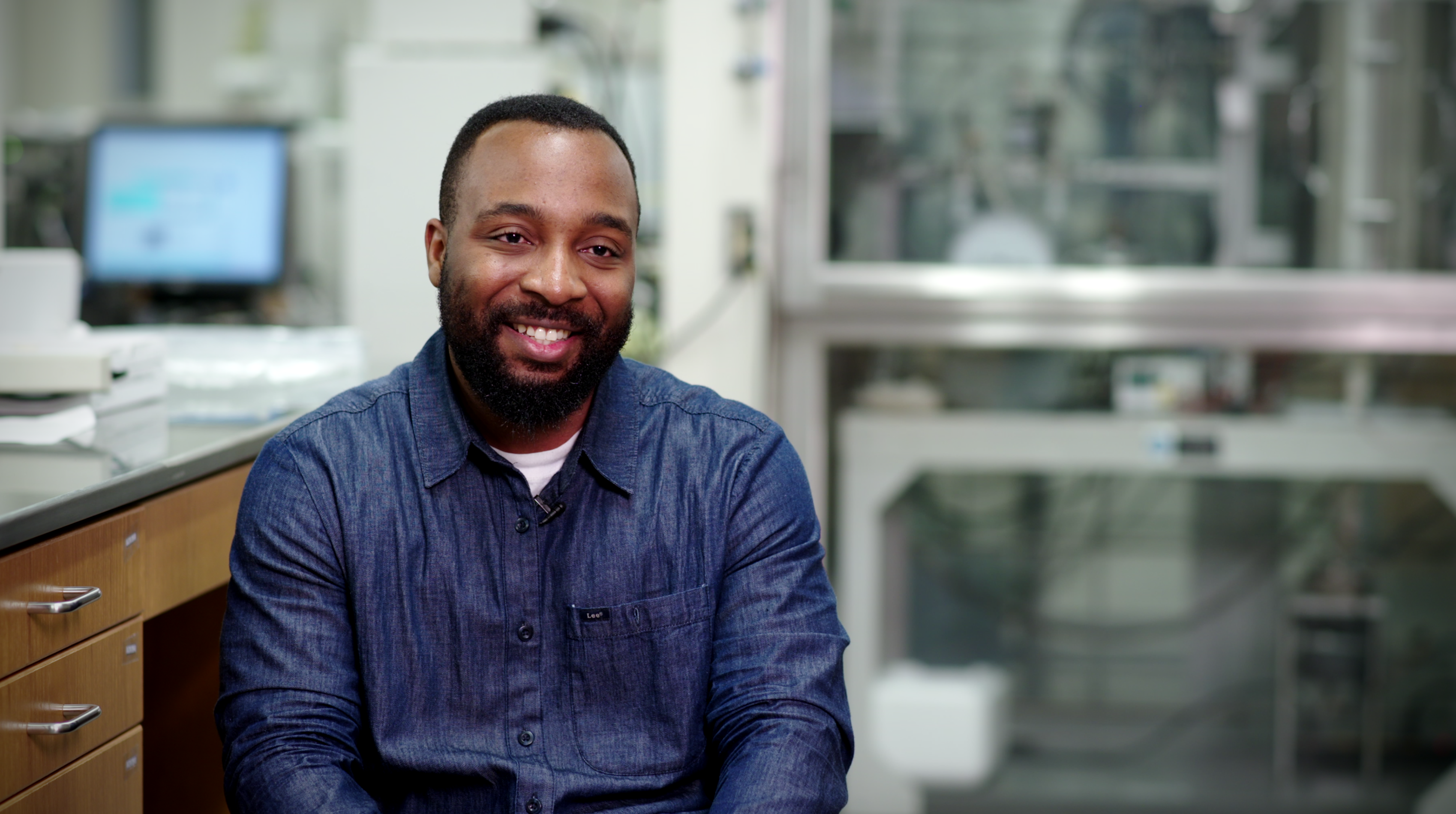 a black man in a blue shirt sits in front of lab machinery and lab tables. he is in focus and the background is not, as if he were being filmed for an interview