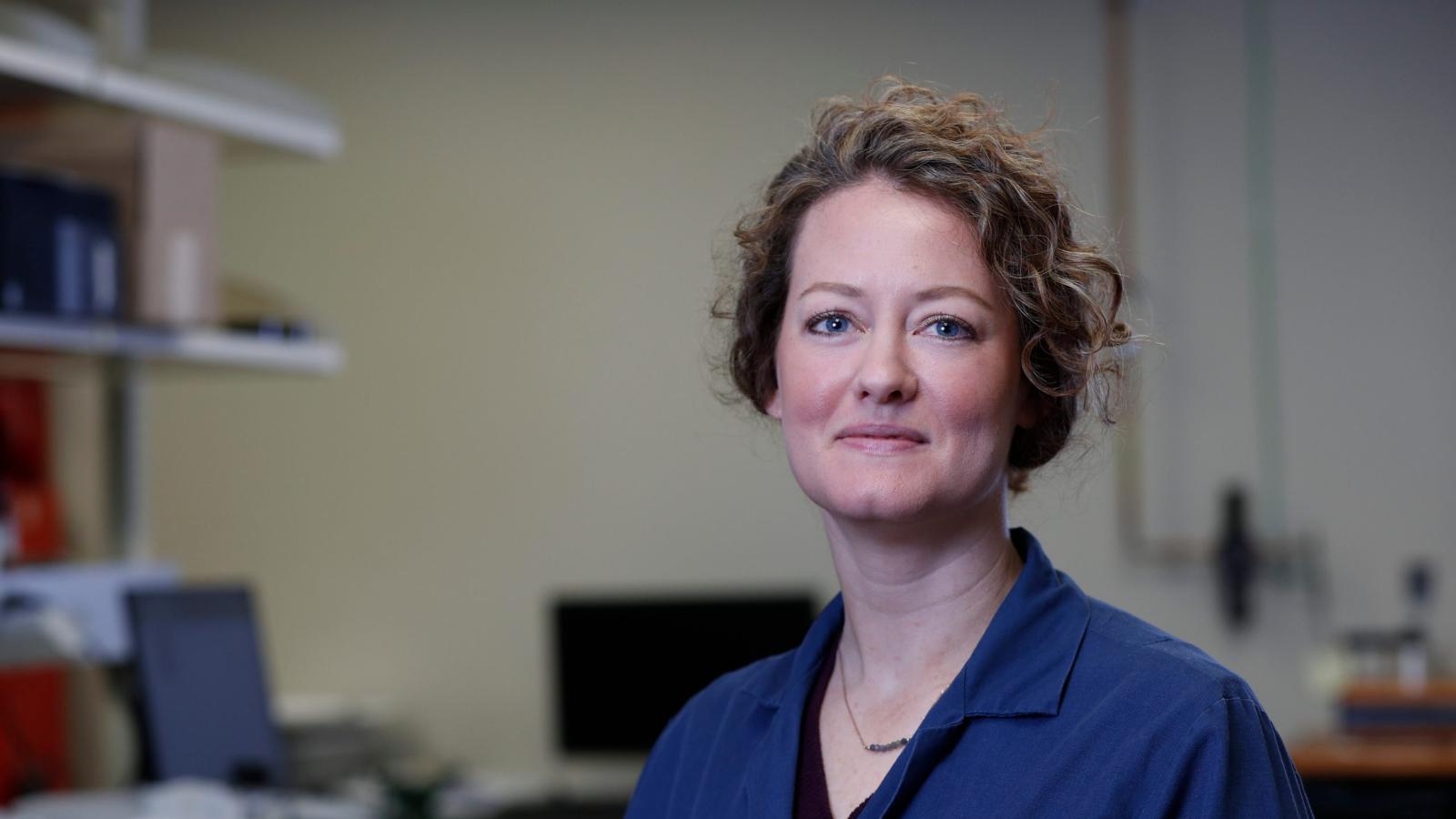 A female researcher stands in an office - the desks are out of focus behind her. She is wearing a blue lab coat and her curly hair is in an updo.
