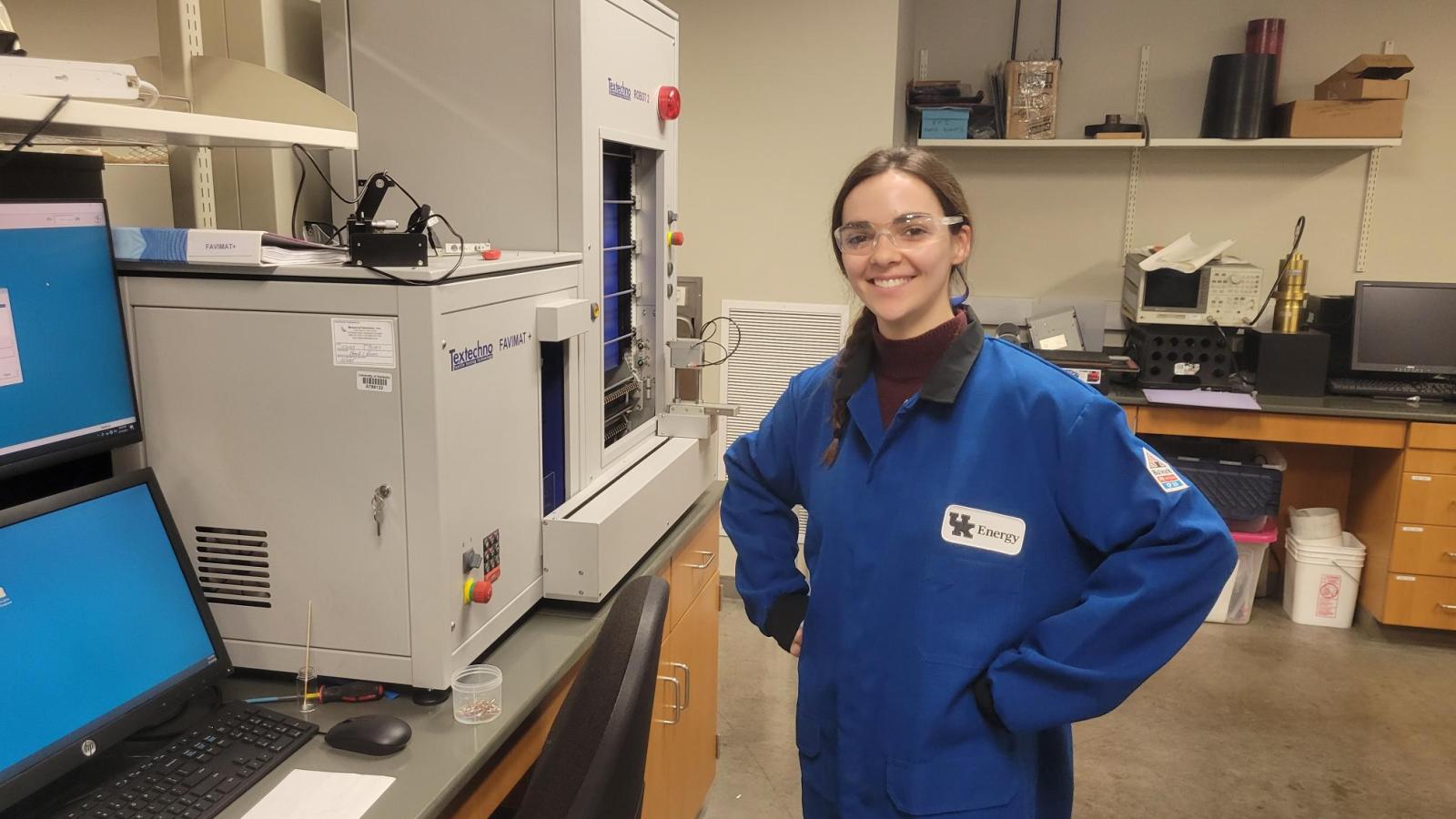 A young woman in lab gear smiles next to analytical instruments