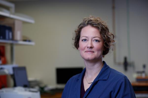 A female researcher stands in an office - the desks are out of focus behind her. She is wearing a blue lab coat and her curly hair is in an updo.