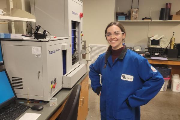 A young woman in lab gear smiles next to analytical instruments