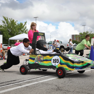 Photo of the beginning of a Gen-EV race. One student pushes a small electric car while another student drives it, preparing to engage the throttle.