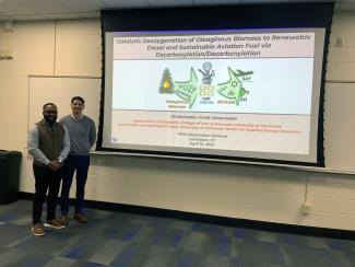 two men, a young black man and a slightly older white man, stand in front of a projector screen with the first slide of a presentation visible