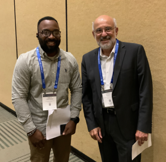 a young black man and an older white man stand next to each other, facing the camera and holding papers