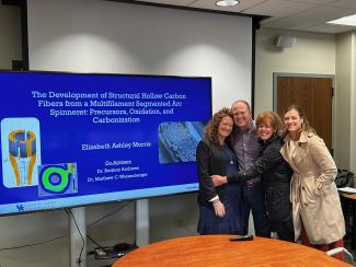 4 people standing in front of a large screen. they are smiling and embracing