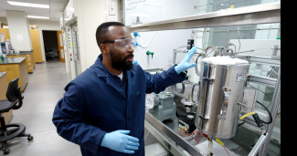 A college-aged black man stands to the left of a piece of complicated lab equipment. He is clearly indicating to the equipment as if explaining it.